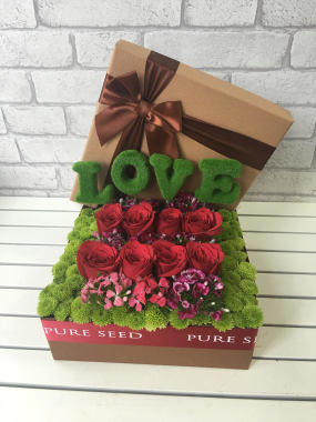 BK540 {Various Size}  -  The lid of a gift box is lifted to reveal a bevy of Roses, decorated with Sweet Williams, embedded in a bed of Pom Poms; the message of LOVE is spelled out in complementary decorative letters.